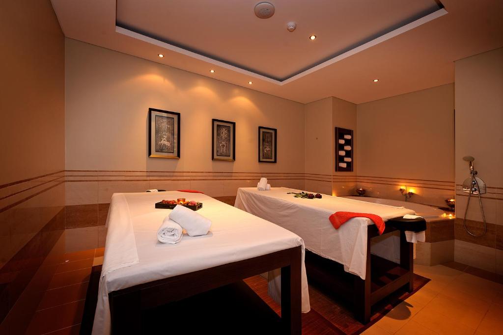 Mierīgā spa telpa ar divām masāžas galdiem, rotātiem ar dvieļiem un ziediem, un siltu apgaismojumu, kas rada relaksējošu atmosfēru.