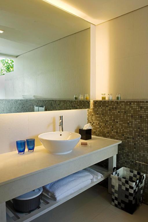 Mūsdienīgs vannas istabas interjers ar elegantu izlietni, divām zilām glāzēm un flīzēm uz sienas. Gaisma un zaļie augi piešķir telpai svaigumu un dzīvīgumu.