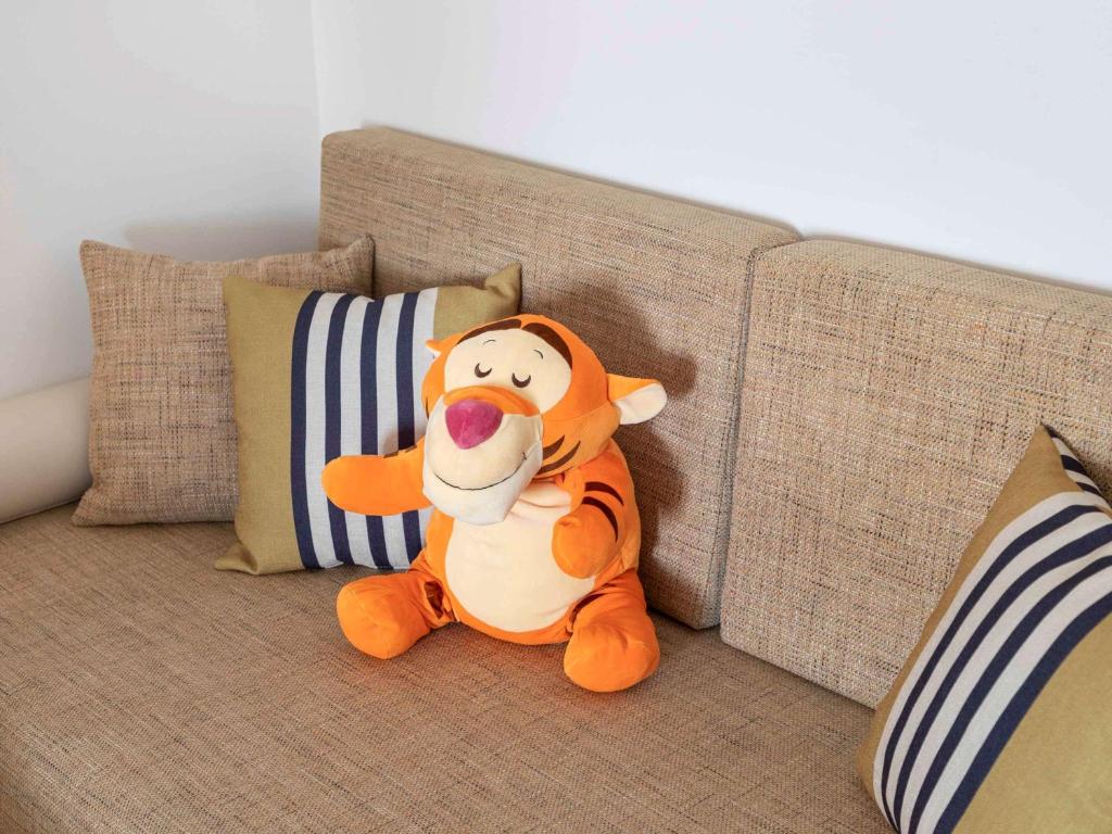 Mīkstā rotaļlieta Tigger sēž uz dīvāna starp spilveniem ar svītrām, radot mājīgu un bērnišķīgu atmosfēru.