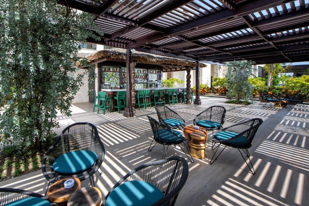 Atvērta terase ar modernām mēbelēm, kurā ir ērtas krēsli un bārs ar dažādiem dzērieniem, ieskauta zaļumiem un saules gaismā veidotiem ēnu rakstiem.