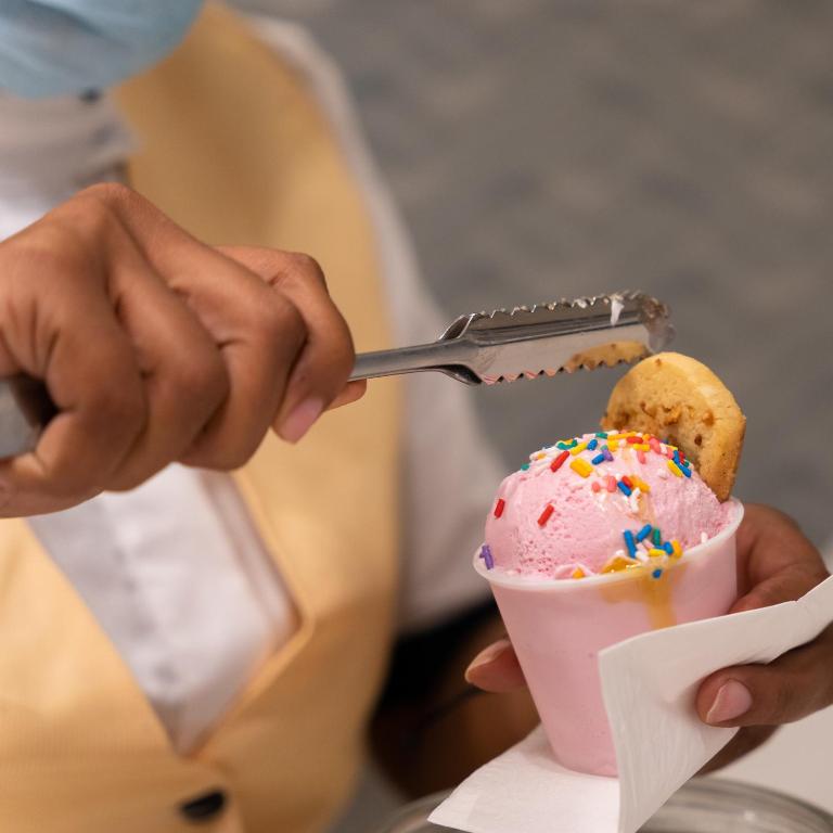 Rokas ar tējkaroti pievieno cepumu krēmveida saldējuma bļodiņai, kas ir rozā krāsā un dekorēta ar konfeti.