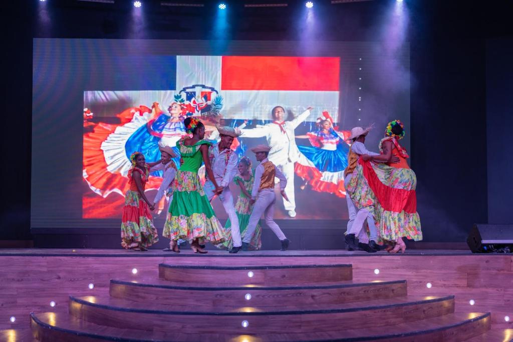 Dejotāju grupa izpilda krāšņu tautas deju uz skatuves, kamēr fona ekrānā redzama līdzīga deju aina. Deju tērpi ir spilgti un krāsaini, radot dzīvīgu atmosfēru.