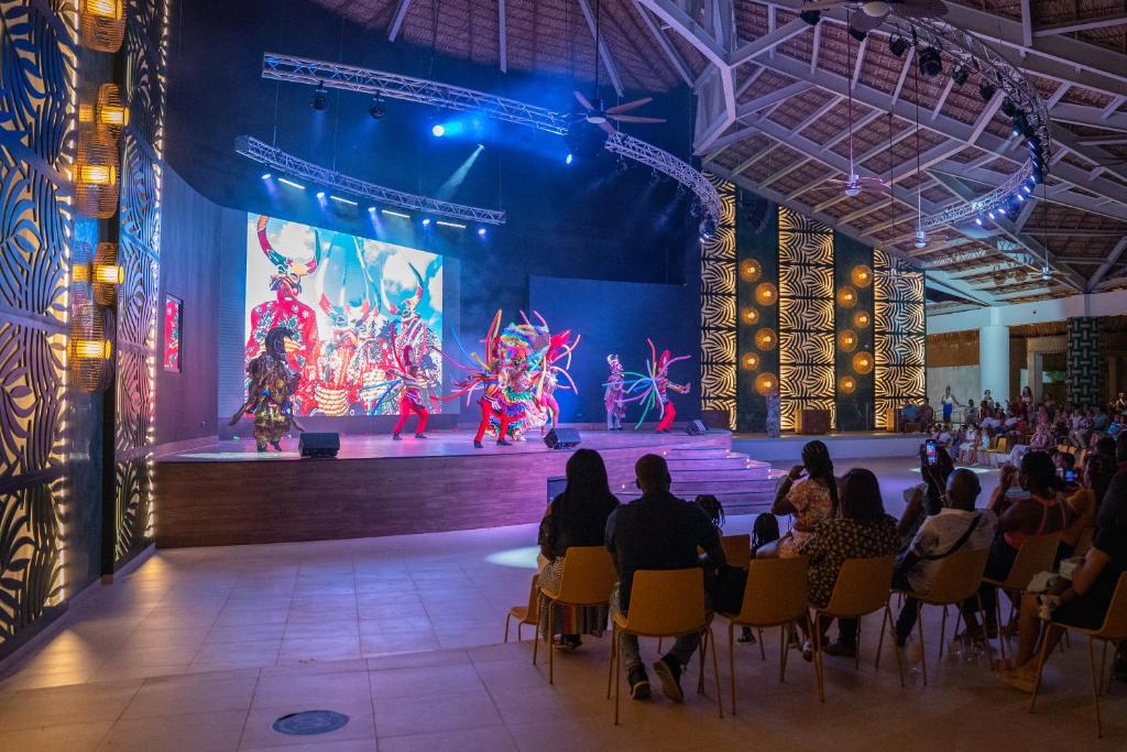 Krāsains kultūras priekšnesums uz skatuves ar dejotājiem, tērptiem tradicionālās drēbēs, un spilgtu gaismu apgaismojumu. Aiz dejotājiem redzama liela ekrāna projekcija, kas piešķir papildus vizuālo efektu.