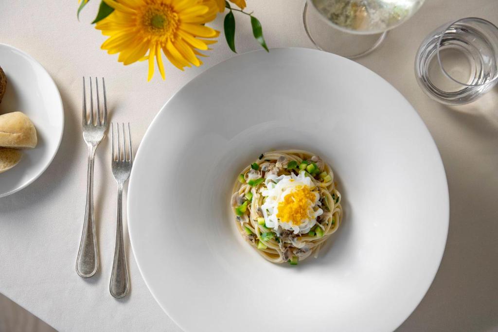 Gardi makaronu ēdiens ar dārzeņiem un olu, pasniegts elegantā baltā traukā. Uz galda redzami arī maizes rullīši un glāze ūdens.