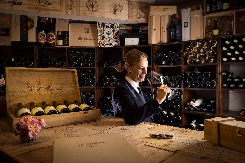 Sieviete, tērpta eleganti, izbauda vīna garšu, turēt glāzi rokās, kamēr fonā redzams plašs vīnu krājums. Uz galda atrodas koka kaste ar vīnu un ziedi.