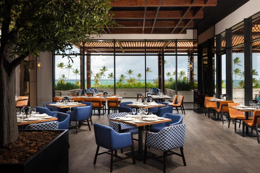 Mūsdienīgs restorāns ar plašām logiem, kas piedāvā skatu uz zilu jūru un palmu kokiem, ar elegantiem galdiem un krēsliem. Interjers apvieno modernus elementus ar dabas akcentiem.