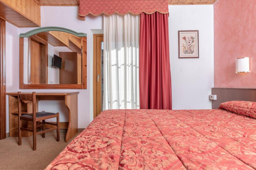 Mūsdienīga viesnīcas istaba ar sarkanu rakstainu gultu, koka mēbelēm un dīvainu spoguli. Aiz loga redzami baltie aizkari un gaišā dienas gaisma.