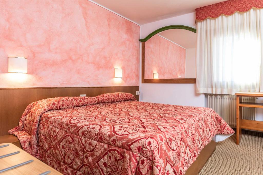 Mājīgi iekārtota guļamistaba ar rozā sienām, sarkanu rakstainu gultas veļu un koka mēbelēm, sniedzot patīkamu atmosfēru atpūtai.