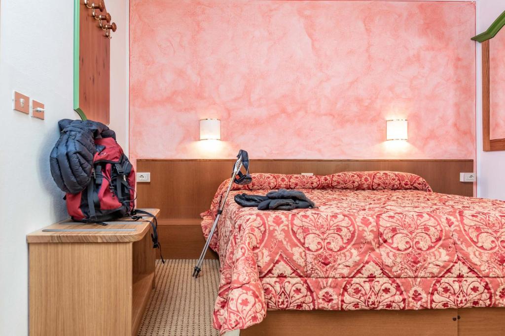 Gultas istaba ar rozā sienām, dekorēta ar ornamenti, uz galda redzama mugursoma un drēbes. Apgaismojums ir maigs, radot mājīgu atmosfēru.