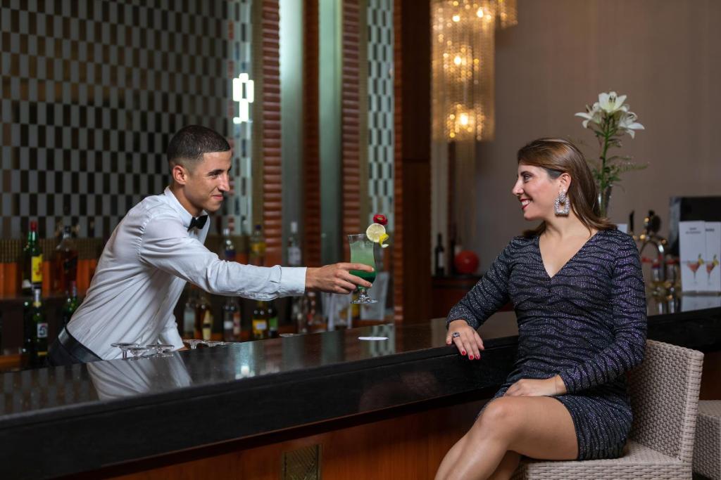 Bārmenis pasniedz dzērienu eleganti ģērbtai sievietei, kas sēž pie bāra. Elegantā atmosfēra un profesionāla apkalpošana rada patīkamu vakara noskaņu.