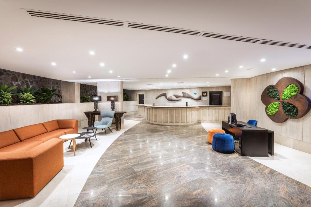 Mūsdienīgs viesnīcas reģistratūras interjers ar stilīgu oranžā dīvāna un dažādu krāsu krēslu kombināciju, kā arī dabīgu augu akcentiem. Apgaismojums rada silti sveiciena atmosfēru.