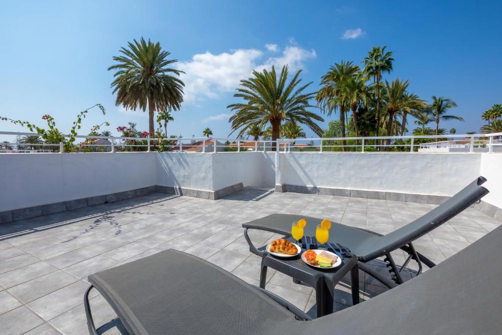 Sauļošanās zona ar divām guļamvietām un galdu, uz kura atrodas brokastu plate ar kruasāniem un dzērieniem, fona veido palmām un skaidra debess.