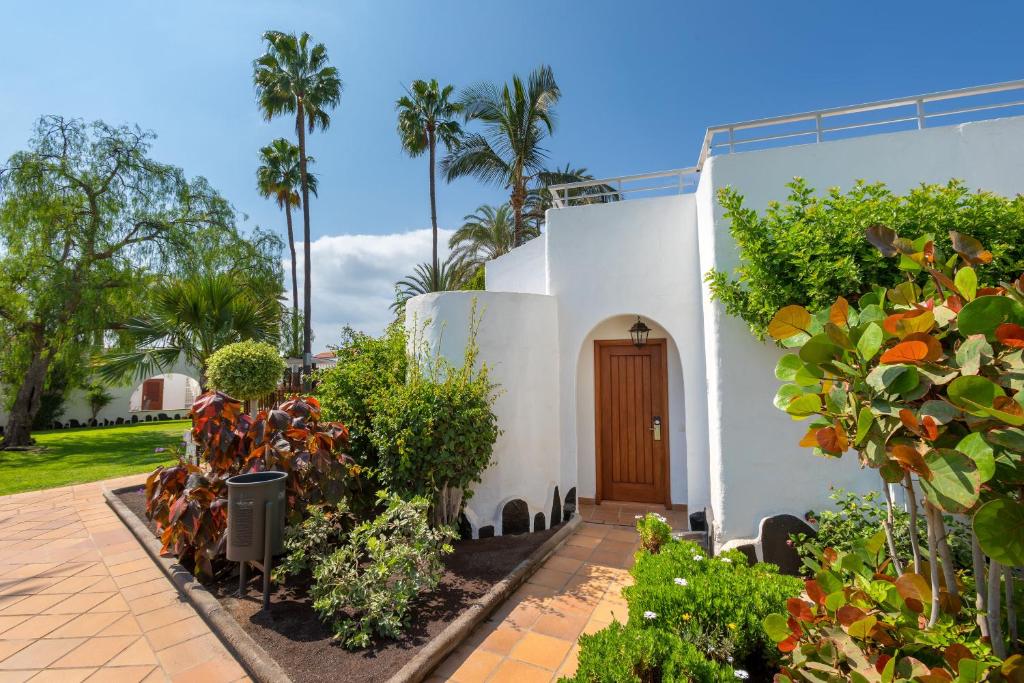 Balta māja ar koka durvīm, ieskauta zaļumiem un palmām, veido patīkamu ainavu. Apmales un ziedi papildina dārzu, radot mierīgu atmosfēru.