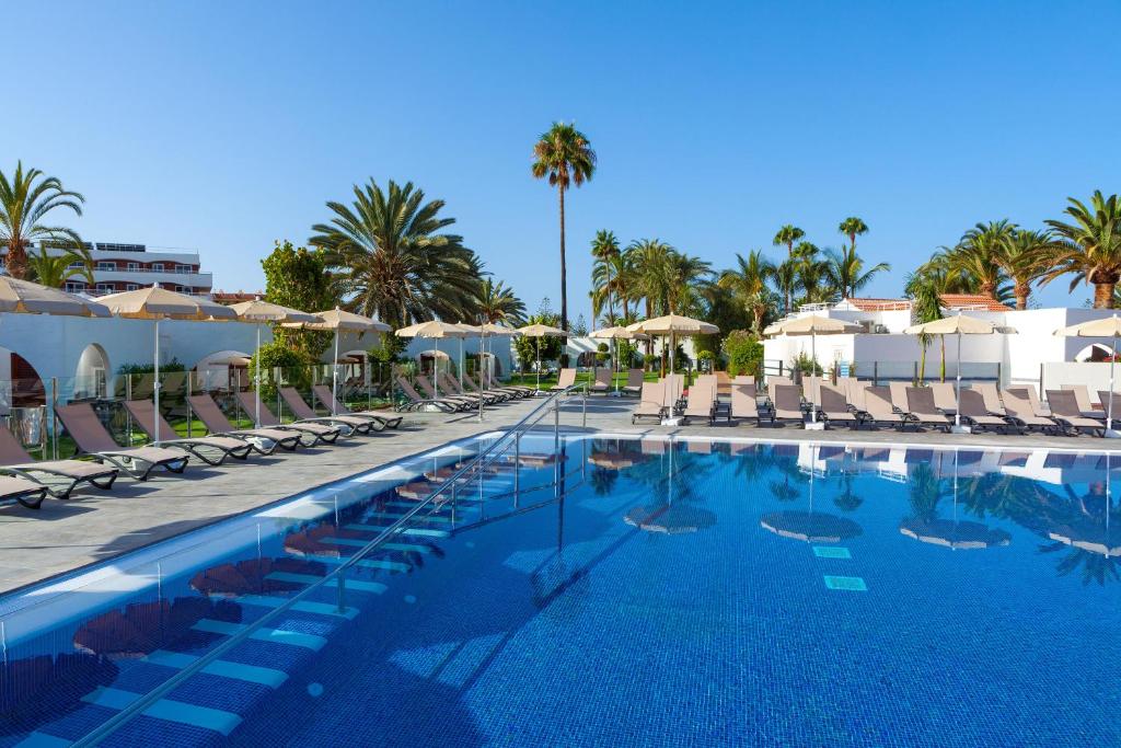 Lieliska viesnīcas baseina zona ar sauļošanās krēsliem un palmu kokiem, nodrošinot relaksējošu atmosfēru saulainā dienā.