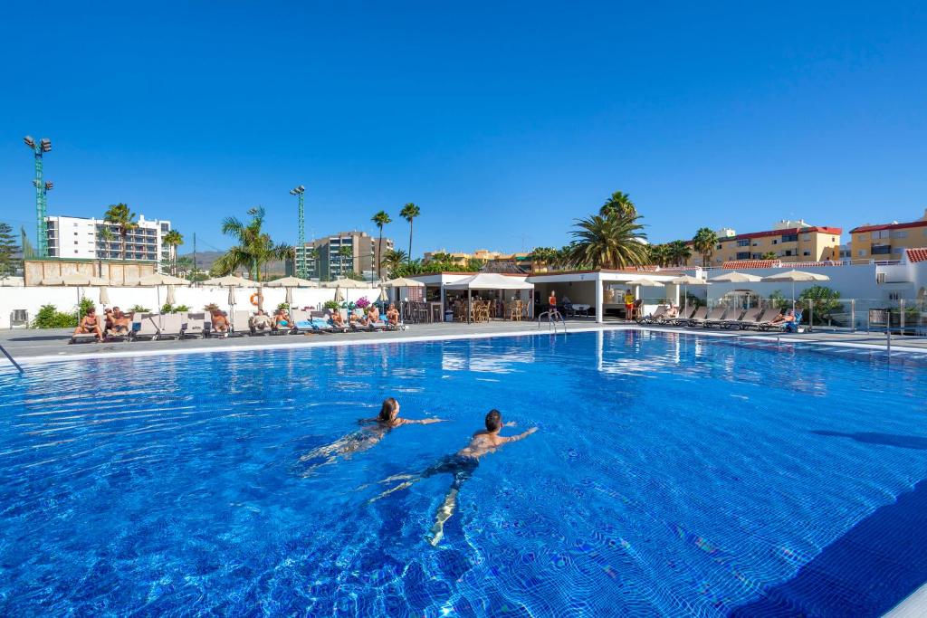 Liels peldbaseins ar skaidru zilu ūdeni, kur divi cilvēki peld, kamēr citi atpūšas blakus. Apkārt baseinam ir palmām un viesnīcu ēkām, radot atpūtas atmosfēru.