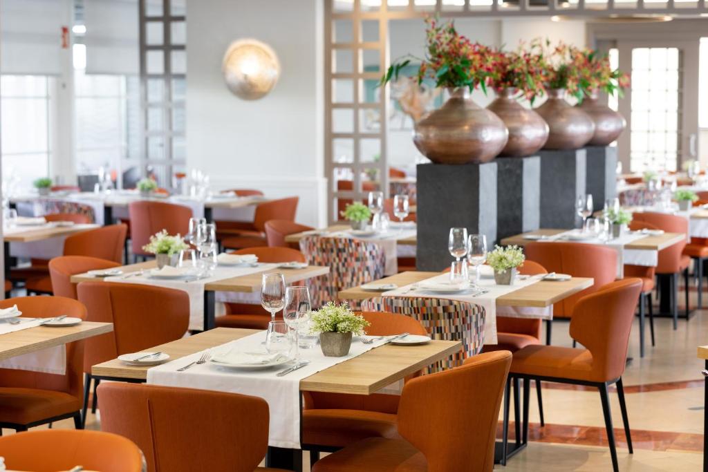 Mūsdienīga restorāna iekārtojums ar oranžām krēslām un eleganti klātiem galdiem, dekorētiem ar ziediem un stilīgiem traukiem. Atmosfēra ir gaumīga un viesmīlīga.