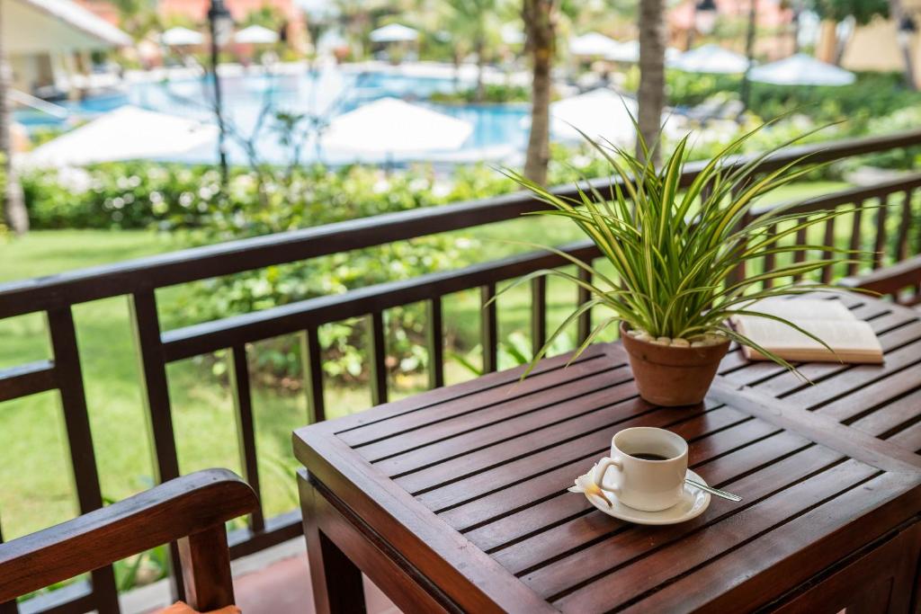 Kafejnīca uz terases ar skatu uz baseinu, kur uz koka galda atrodas kafijas tasīte, šķīvis ar cepumu un puķu pods ar zaļo augu.