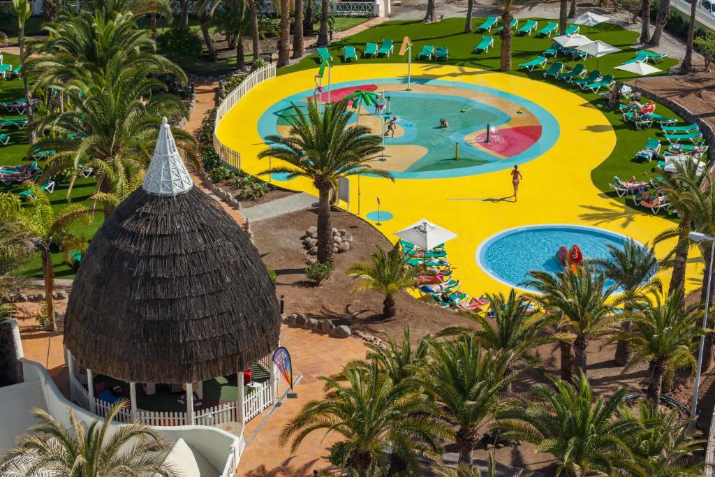 Krāsains ūdens parks ar bērnu rotaļu zonu un peldbaseinu, ieskauts palmām un sauļošanās krēsliem. Ideāla vieta atpūtai un izklaidei.