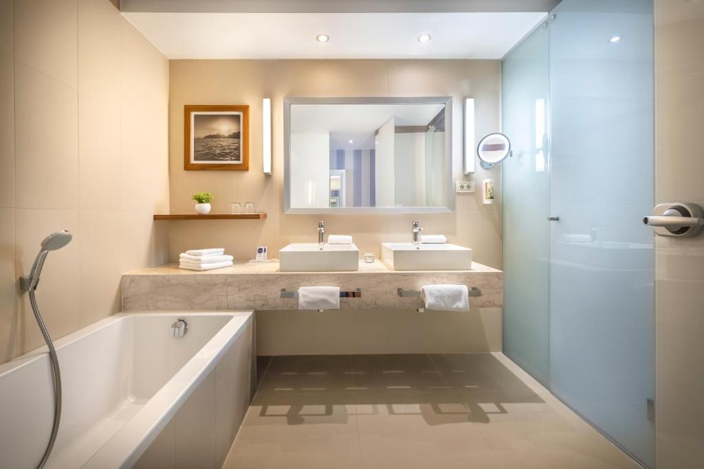 Mūsdienīgs vannas istabas interjers ar diviem izlietnēm, dušu un vannu, papildināts ar dekoratīvu attēlu un zaļumu.