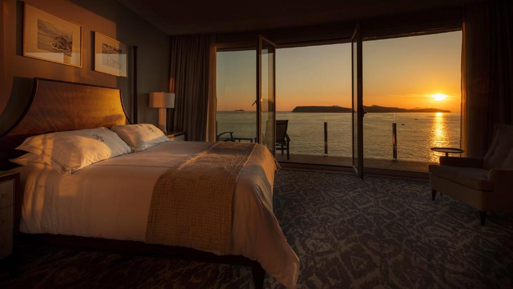 Greznas viesnīcas numurs ar skatu uz saulrietu pār jūru, kur ir ērta gulta un plaši logi. Dabiskais apgaismojums rada mierīgu atmosfēru.