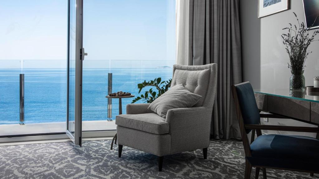 Mūsdienīgs viesnīcas numurs ar skatu uz jūru, kurā ir ērta pelēka krēsla, stikla durvju, kas ved uz balkonu, un dekoratīvas augu detaļas.
