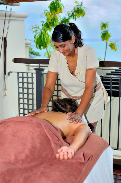 Masāžas sesija spa vidē, kur masiere sniedz relaksējošu masāžu klientam. Apkārt ir tropiska vide ar zaļām palmām un jūras skatu.