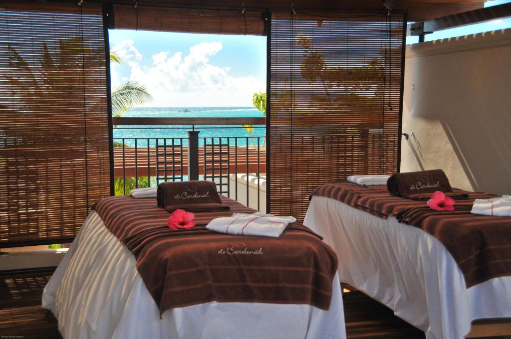 Relaksējoša spa telpa ar divām masāžas galdiem, rotātiem ar dvieļiem un ziediem, logi sniedz skatu uz jūru un palmu ainavu.