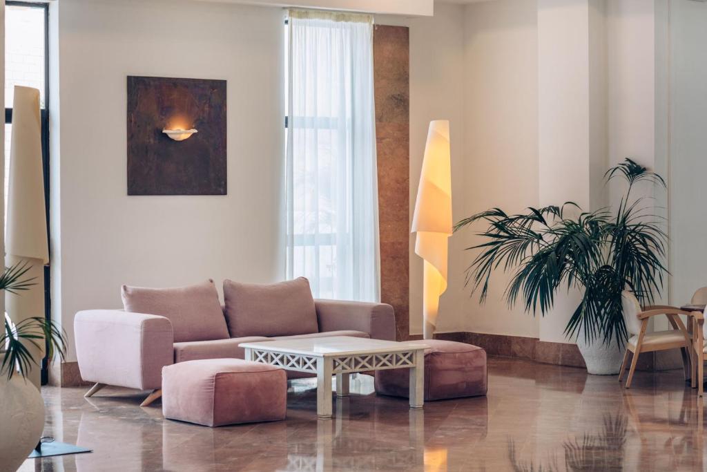 Mūsdienīgs viesnīcas viesistabas interjers ar rozā dīvānu, balto kafijas galdiņu un sulīgu palmu stūri. Gaismas ķermenis un lieli logi piepilda telpu ar dabu un gaismu.