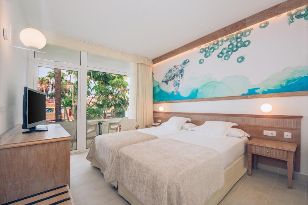 Moderns viesnīcas numurs ar divām gultām, palīgtabulu un televizoru, kas sniedz skatu uz zaļām palmām caur lielajiem logiem. Sienā ir māksliniecīgs attēls ar jūras bruņurupuci.