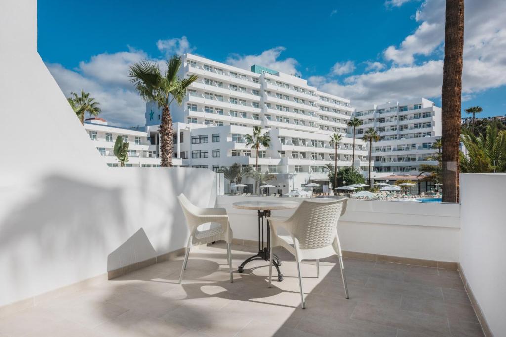 Mūsdienīgs viesnīcas balkons ar galdu un krēsliem, skats uz baseinu un apkārtējiem palmām. Saulains laiks un skaidras debesis papildina relaksējošo atmosfēru.