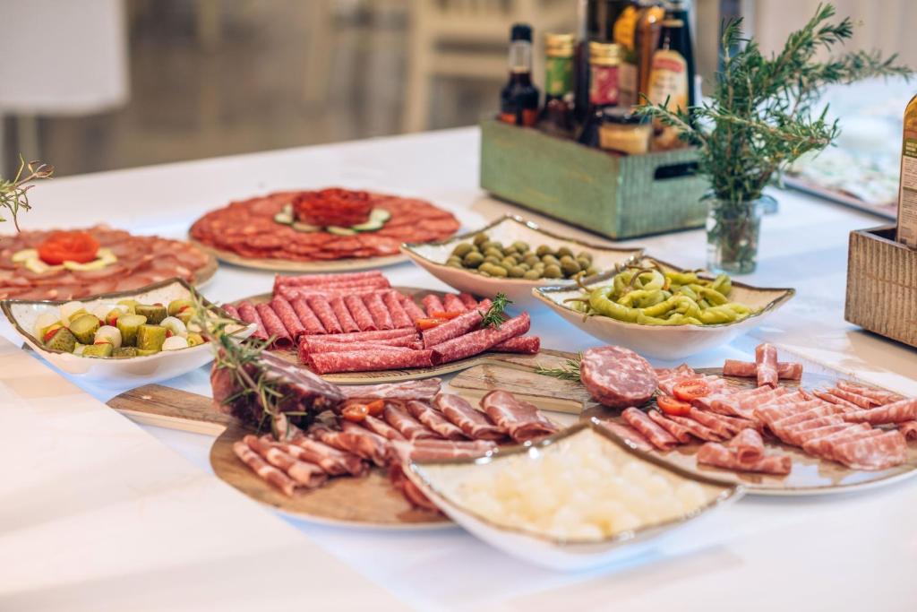 Dažādu gaļas un siera uzkodu assortiments uz baltas galda virsmas, papildināts ar marinētiem dārzeņiem un garšaugiem.
