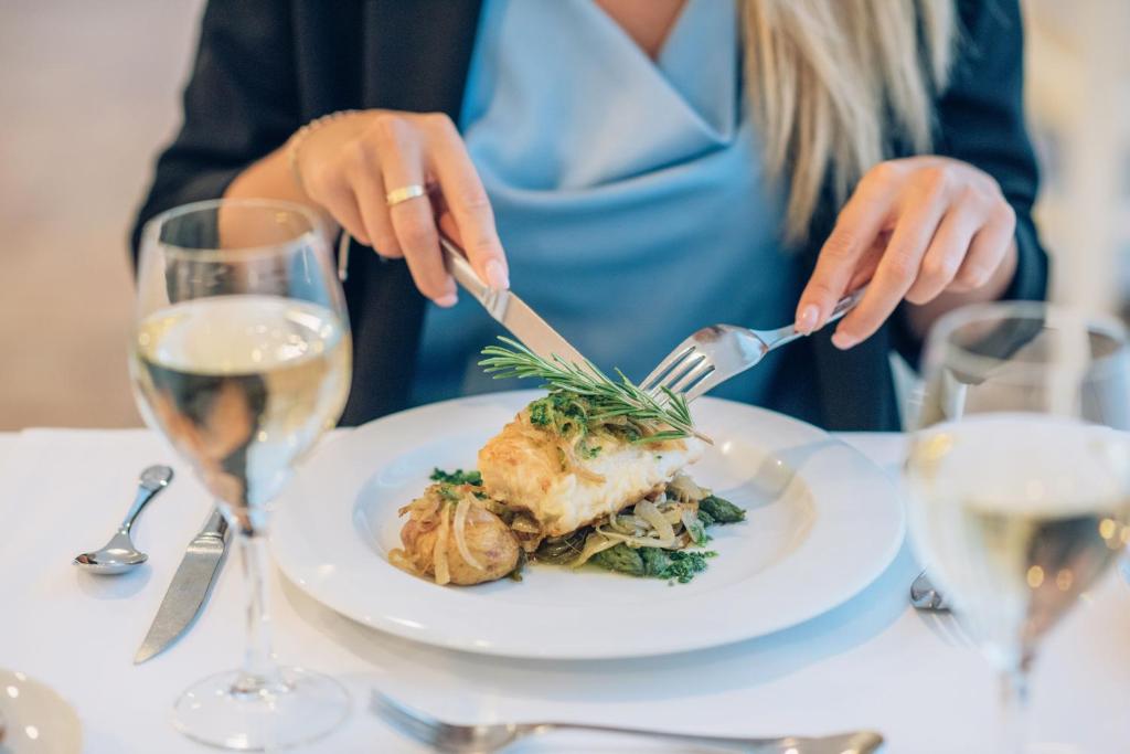 Eleganta maltīte restorānā ar ceptu zivi un dārzeņiem, sieviete ar dakšiņu un nazi gatavojas ēst. Uz galda ir glāze baltā vīna un galda piederumi.