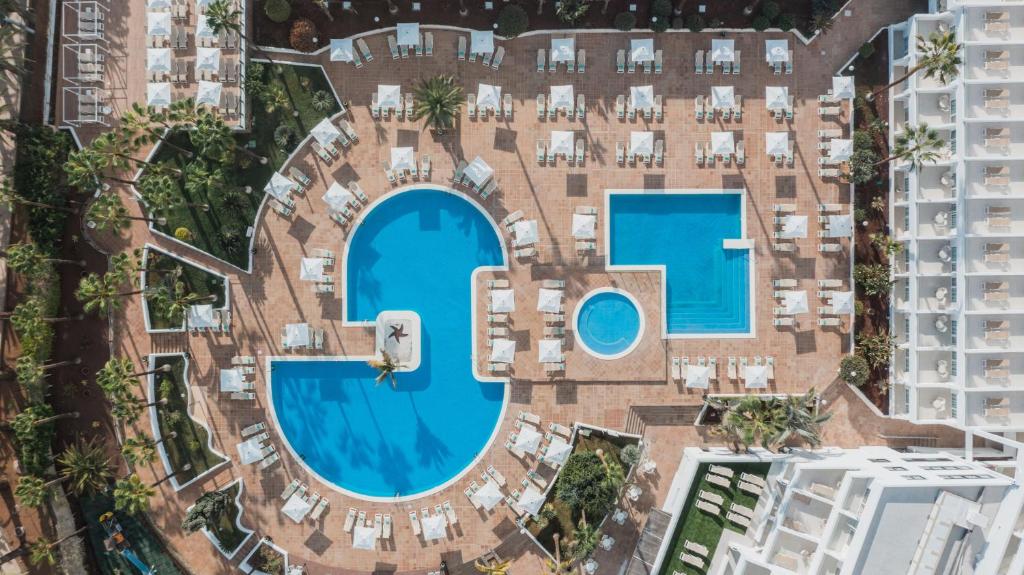 Viesnīcas baseina un atpūtas zonas panorāmas skats, kurā redzami divi baseini un saulessargu rindu. Apkārtne ir zaļa ar palmām un ērtām atpūtas vietām.