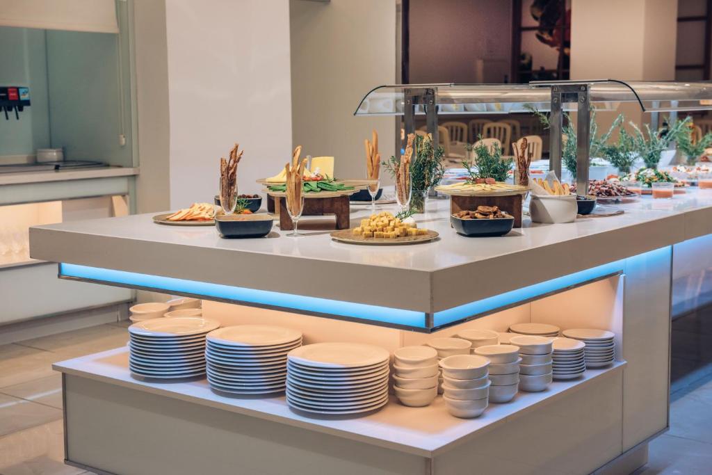 Brokastu bufete ar dažādiem ēdieniem, tostarp uzkodām un saldumiem, modernā un eleganti iekārtotā telpā. Galda apakšā redzamas balta trauki.