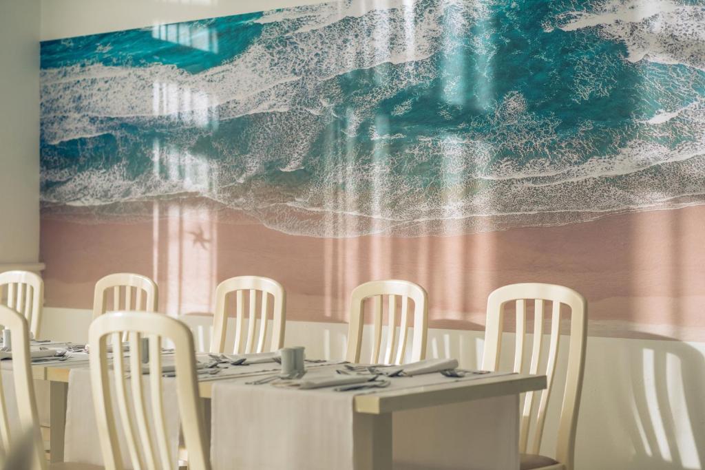 Restorāna interjers ar pludmales ainavu uz sienas, kurā redzami balti galdi un krēsli.