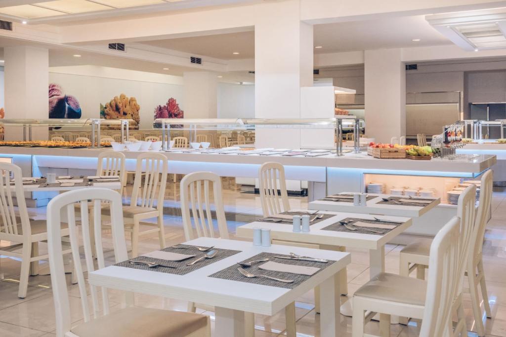 Mūsdienīgs restorāns ar baltām mēbelēm un plašu bufetes galdu, kurā redzami dažādi ēdieni. Interjerā ir gaišas krāsas un dekorācijas, kas rada patīkamu atmosfēru.