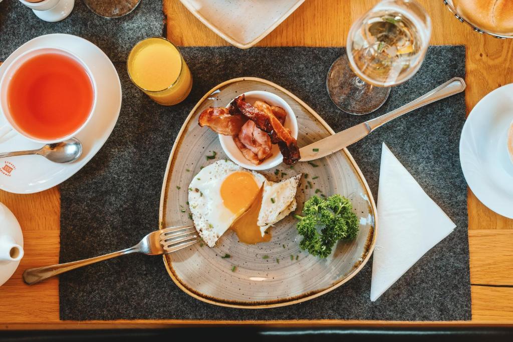 Broka piegāde ar ceptiem olu dzeltenumiem, bekona šķēlītēm un svaigu pētersīļu lapām, kopā ar tēju un sulu, izvietota uz eleganta galda.