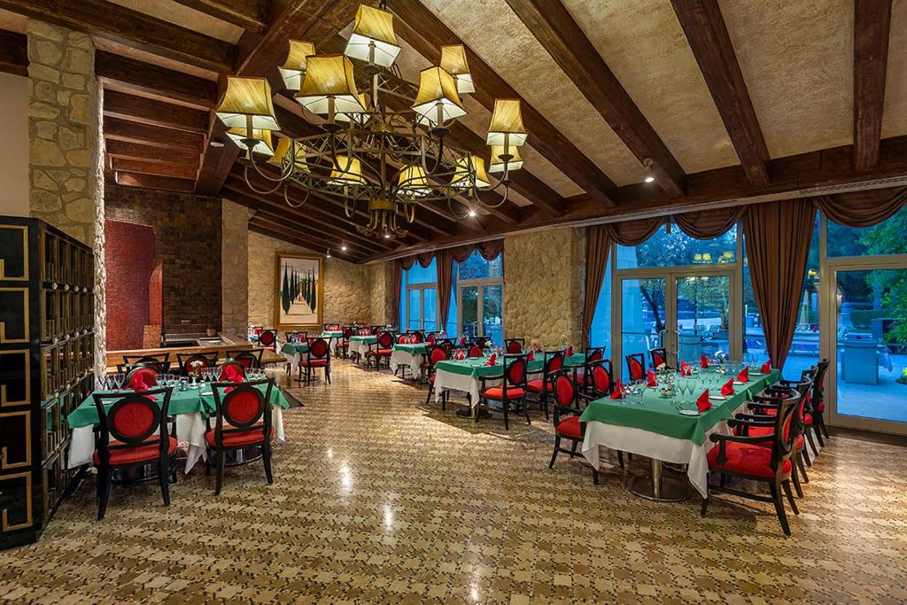 Elegants restorāns ar koka sijām un akmens sienām, kurā ir zaļi galdi ar sarkaniem salvetēm. Gaismas lustra piešķir siltu atmosfēru, logi paver skatu uz dārzu.