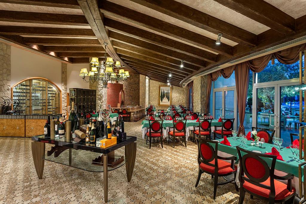 Elegants restorāns ar sarkaniem krēsliem un zaļiem galdautiem, kas izceļas ar koka sijām un romantisku apgaismojumu. Uz galda redzami dažādi dzērieni, kas papildina atmosfēru.