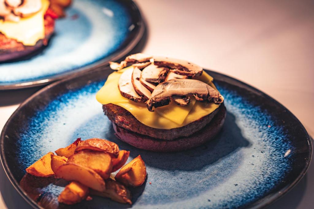 Saldēta burgera pamatne ar cheddar sieru un sēnēm, pasniegta uz zila šķīvja kopā ar ceptiem kartupeļiem.