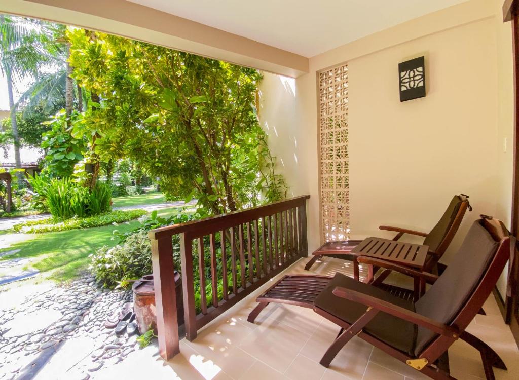 Mierīga balkona zona ar divām koka atpūtas krēslām, no kuras paveras skats uz zaļojošu dārzu un palmu kokiem. Ideāla vieta relaksācijai un dabas baudīšanai.