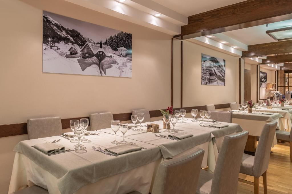 Elegants restorāna interjers ar baltām galdautām un galdiem, kas sagatavoti viesu uzņemšanai. Sienās redzami melnbalti kalnu attēli, radot mierīgu atmosfēru.