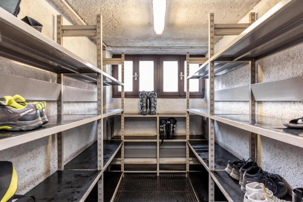 Tukšs skapis ar metāla plauktiem un dažām apaviem, kas novietoti uz grīdas. Logi nodrošina dabisko apgaismojumu, radot gaišu un sakārtotu vidi.