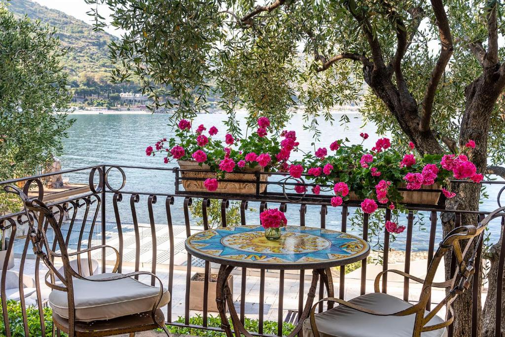 Balkons ar skatu uz ūdeni, dekorēts ar ziediem un krāsainu galdu, radot mierīgu un relaksējošu atmosfēru. Ideāla vieta atpūtai un baudīšanai.