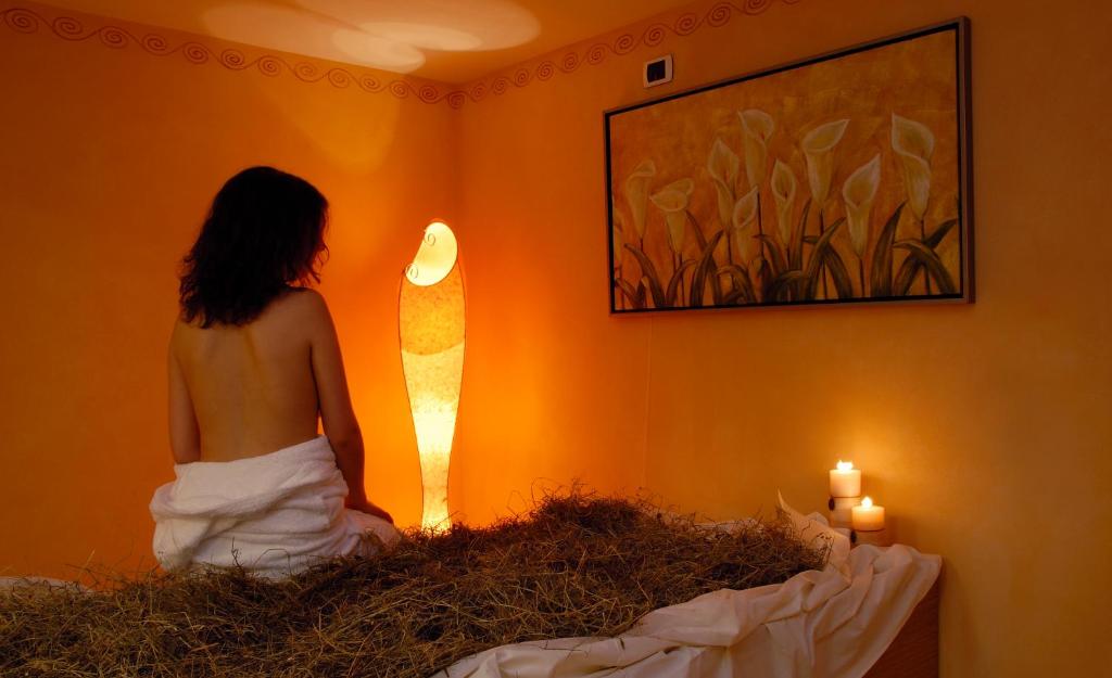 Mierīga spa atmosfēra ar apgaismojumu un sveču gaismu, kur sieviete atpūšas uz gultas, kas klāta ar zālēm. Sienā ir glezna ar ziediem, radot relaksējošu vidi.