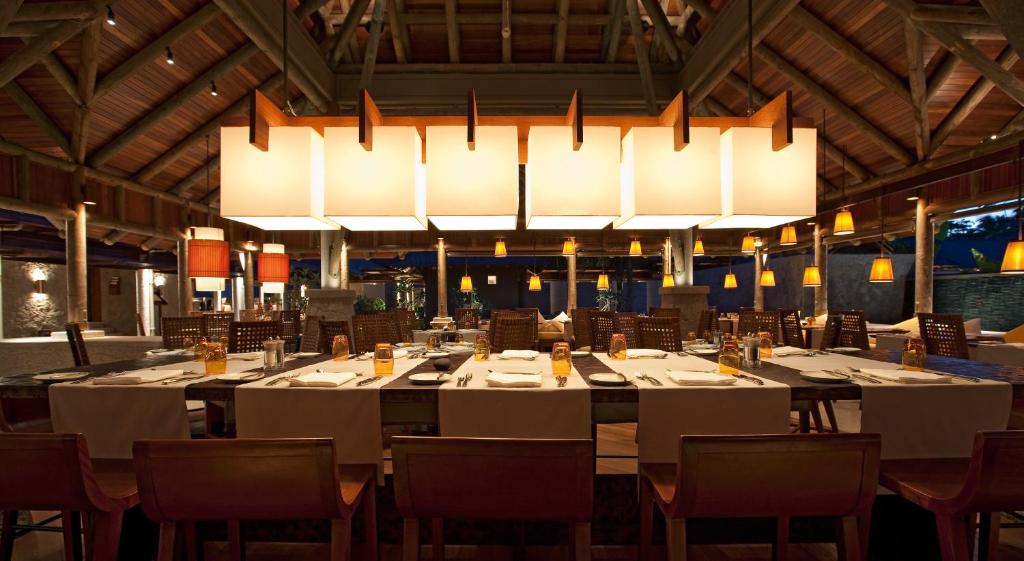 Mūsdienīga restorāna interjers ar koka konstrukcijām un stilīgu apgaismojumu, kas rada siltu un aicinošu atmosfēru. Galdi ir sakārtoti ar galda piederumiem un dekoriem, gatavi viesu ierašanās brīdim.