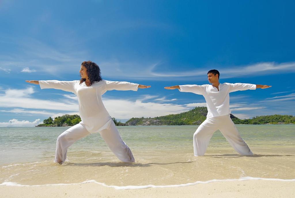 Pāris, tērpies baltos apģērbos, veic jogas pozu pludmalē ar skaistu zilo debesi un jūru fonā.