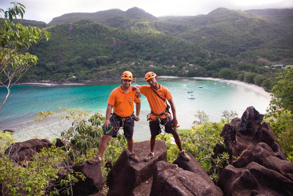 Divi kalnu kāpēji pozē uz akmeņiem ar skaistu pludmali un zilo ūdeni fona. Abiem ir oranži krekli un drošības aprīkojums, kas liecina par piedzīvojumu.