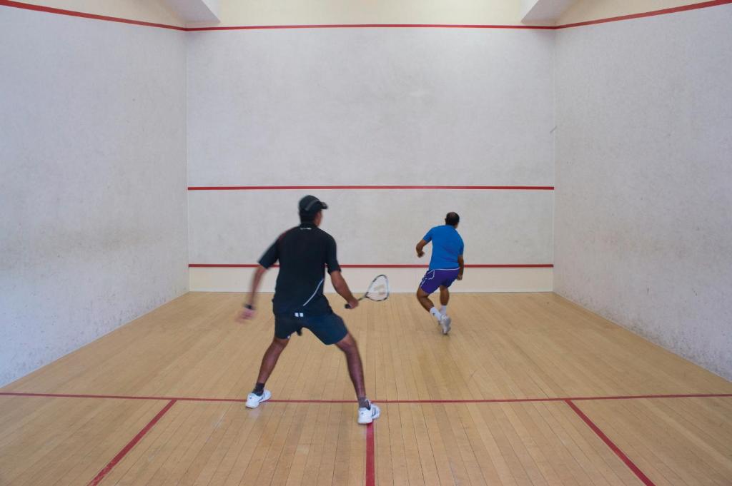 Divi spēlētāji spēlē skvošu, aktīvi kustoties laukumā ar koka grīdu un baltām sienām.