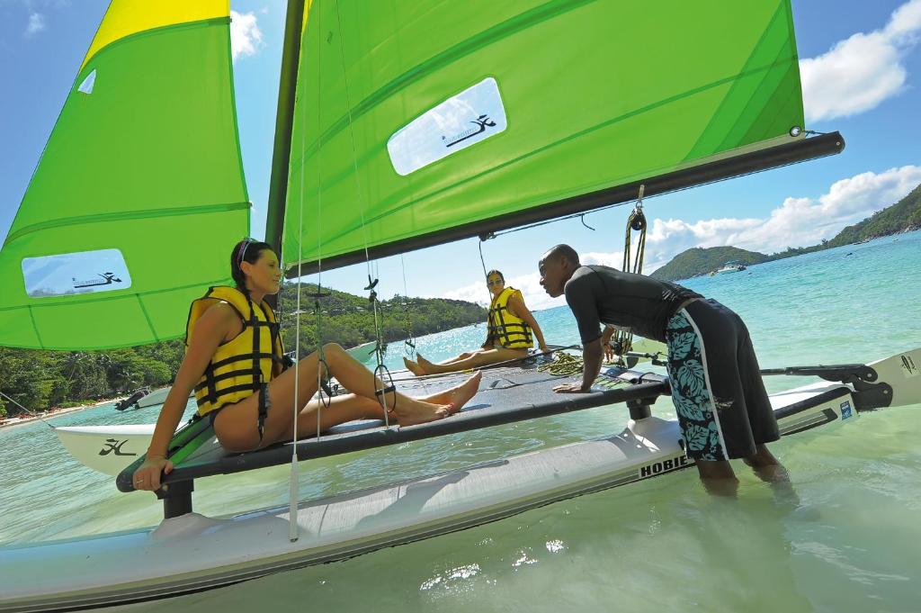 Divi cilvēki relaksējas uz katamarāna ar zaļiem burām, kamēr trešais cilvēks stāv ūdenī blakus, sniedzot norādījumus. Apkārt ir skaidrs, zils ūdens un zaļš ainava.